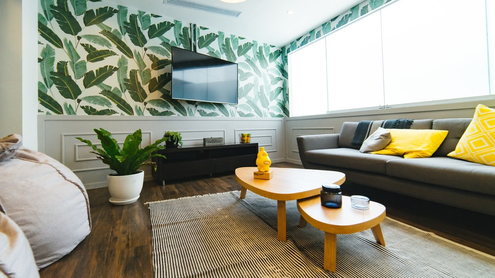 A Living Room Featuring a Gray Custom Sofa with Throw Pillows, Wooden Tables, Wall-mounted TV, and a Wallpaper with a Leafy Pattern 